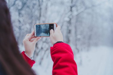 Kız kış aylarında ormanda bir smartphone cep telefonu ile fotoğraf çekmek. Ormanda yürüyen kadın