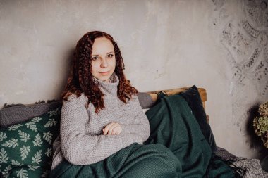 Bir genç kız yatakta oturmuş bir kazaklı bir battaniyeye huddled. Sıcak kazak giymiş kadın sokak stil giyim. Temiz cilt, uzun saç