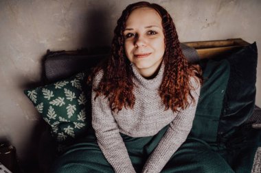 Bir genç kız yatakta oturmuş bir kazaklı bir battaniyeye huddled. Sıcak kazak giymiş kadın sokak stil giyim. Temiz cilt, uzun saç