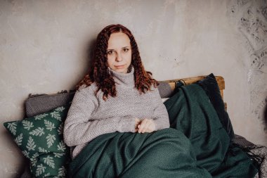 Bir genç kız yatakta oturmuş bir kazaklı bir battaniyeye huddled. Sıcak kazak giymiş kadın sokak stil giyim. Temiz cilt, uzun saç