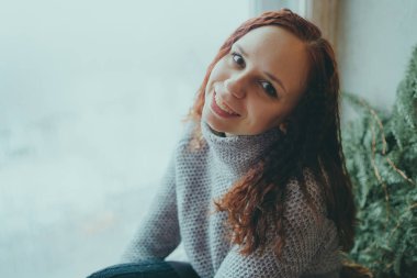 Genç bir kız bir kazaklı pencerenin önünde poz. Sıcak kazak giymiş kadın sokak stil giyim. Temiz cilt, uzun saç