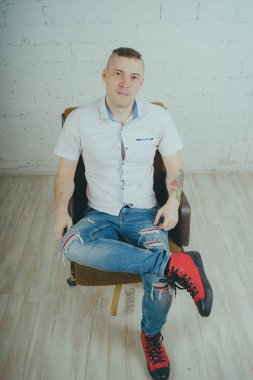 Duygusal ofis çalışanı dövmeli, beyaz duvara karşı bir sandalye üzerinde oturan adam. Bir kravat adam kot pantolon ve beyaz gömlek giymiş.