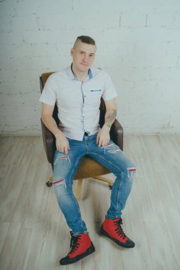 Duygusal ofis çalışanı dövmeli, beyaz duvara karşı bir sandalye üzerinde oturan adam. Bir kravat adam kot pantolon ve beyaz gömlek giymiş.