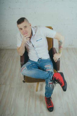 Duygusal ofis çalışanı dövmeli, beyaz duvara karşı bir sandalye üzerinde oturan adam. Bir kravat adam kot pantolon ve beyaz gömlek giymiş.