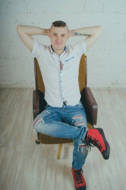 Duygusal ofis çalışanı dövmeli, beyaz duvara karşı bir sandalye üzerinde oturan adam. Bir kravat adam kot pantolon ve beyaz gömlek giymiş.