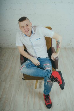 Duygusal ofis çalışanı dövmeli, beyaz duvara karşı bir sandalye üzerinde oturan adam. Bir kravat adam kot pantolon ve beyaz gömlek giymiş.