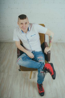 Duygusal ofis çalışanı dövmeli, beyaz duvara karşı bir sandalye üzerinde oturan adam. Bir kravat adam kot pantolon ve beyaz gömlek giymiş.