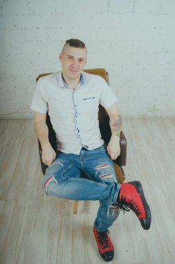 Duygusal ofis çalışanı dövmeli, beyaz duvara karşı bir sandalye üzerinde oturan adam. Bir kravat adam kot pantolon ve beyaz gömlek giymiş.