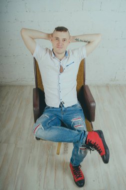 Duygusal ofis çalışanı dövmeli, beyaz duvara karşı bir sandalye üzerinde oturan adam. Bir kravat adam kot pantolon ve beyaz gömlek giymiş.