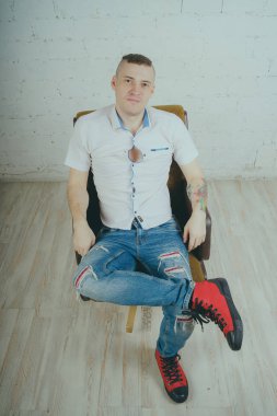 Duygusal ofis çalışanı dövmeli, beyaz duvara karşı bir sandalye üzerinde oturan adam. Bir kravat adam kot pantolon ve beyaz gömlek giymiş.
