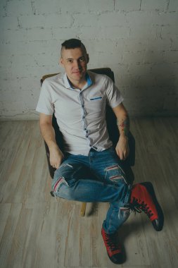 Duygusal ofis çalışanı dövmeli, beyaz duvara karşı bir sandalye üzerinde oturan adam. Bir kravat adam kot pantolon ve beyaz gömlek giymiş.