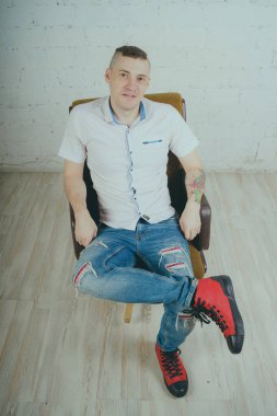 Duygusal ofis çalışanı dövmeli, beyaz duvara karşı bir sandalye üzerinde oturan adam. Bir kravat adam kot pantolon ve beyaz gömlek giymiş.