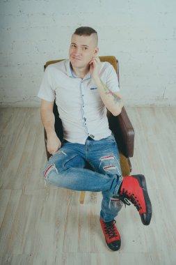 Duygusal ofis çalışanı dövmeli, beyaz duvara karşı bir sandalye üzerinde oturan adam. Bir kravat adam kot pantolon ve beyaz gömlek giymiş.