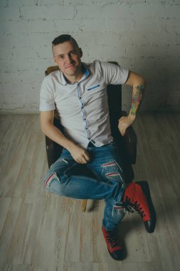 Duygusal ofis çalışanı dövmeli, beyaz duvara karşı bir sandalye üzerinde oturan adam. Bir kravat adam kot pantolon ve beyaz gömlek giymiş.