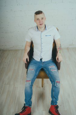 Duygusal ofis çalışanı dövmeli, beyaz duvara karşı bir sandalye üzerinde oturan adam. Bir kravat adam kot pantolon ve beyaz gömlek giymiş.