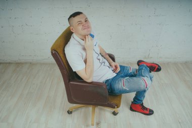 Duygusal ofis çalışanı dövmeli, beyaz duvara karşı bir sandalye üzerinde oturan adam. Bir kravat adam kot pantolon ve beyaz gömlek giymiş.