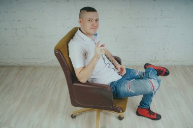 Duygusal ofis çalışanı dövmeli, beyaz duvara karşı bir sandalye üzerinde oturan adam. Bir kravat adam kot pantolon ve beyaz gömlek giymiş.