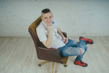 Duygusal ofis çalışanı dövmeli, beyaz duvara karşı bir sandalye üzerinde oturan adam. Bir kravat adam kot pantolon ve beyaz gömlek giymiş.