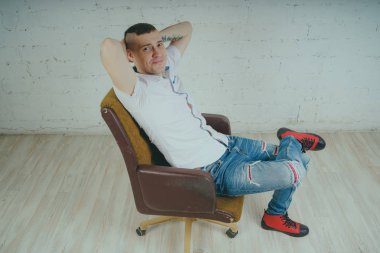 Duygusal ofis çalışanı dövmeli, beyaz duvara karşı bir sandalye üzerinde oturan adam. Bir kravat adam kot pantolon ve beyaz gömlek giymiş.