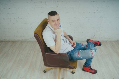 Duygusal ofis çalışanı dövmeli, beyaz duvara karşı bir sandalye üzerinde oturan adam. Bir kravat adam kot pantolon ve beyaz gömlek giymiş.