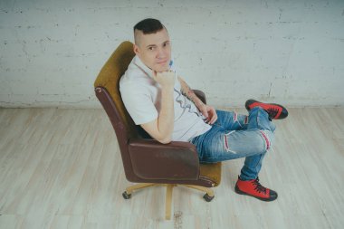 Duygusal ofis çalışanı dövmeli, beyaz duvara karşı bir sandalye üzerinde oturan adam. Bir kravat adam kot pantolon ve beyaz gömlek giymiş.