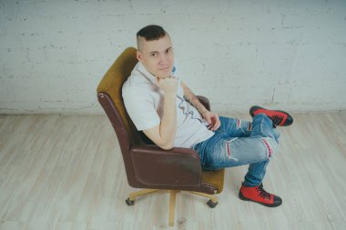 Duygusal ofis çalışanı dövmeli, beyaz duvara karşı bir sandalye üzerinde oturan adam. Bir kravat adam kot pantolon ve beyaz gömlek giymiş.