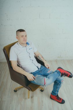 Duygusal ofis çalışanı dövmeli, beyaz duvara karşı bir sandalye üzerinde oturan adam. Bir kravat adam kot pantolon ve beyaz gömlek giymiş.