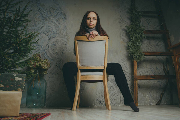 A young girl in a sweater posing against a gray wall. Woman dressed in warm sweater, street style clothing. Clean skin, long hair