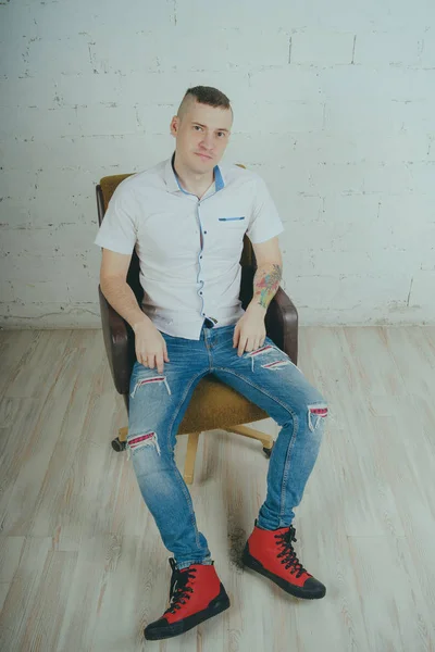 Duygusal ofis çalışanı dövmeli, beyaz duvara karşı bir sandalye üzerinde oturan adam. Bir kravat adam kot pantolon ve beyaz gömlek giymiş.