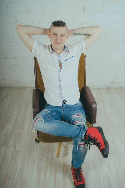 Duygusal ofis çalışanı dövmeli, beyaz duvara karşı bir sandalye üzerinde oturan adam. Bir kravat adam kot pantolon ve beyaz gömlek giymiş.