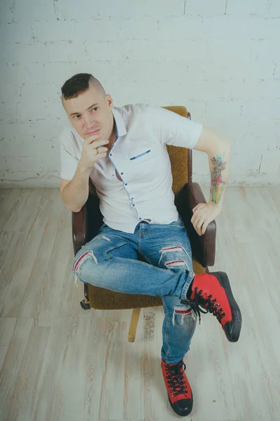 Duygusal ofis çalışanı dövmeli, beyaz duvara karşı bir sandalye üzerinde oturan adam. Bir kravat adam kot pantolon ve beyaz gömlek giymiş.