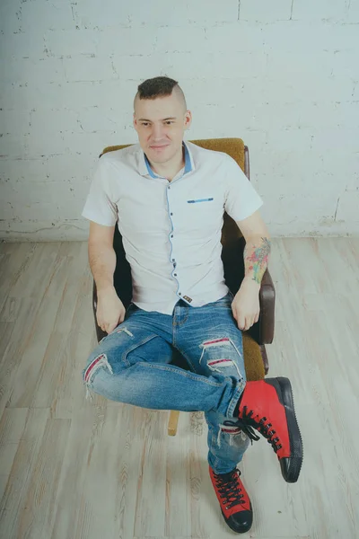 Duygusal ofis çalışanı dövmeli, beyaz duvara karşı bir sandalye üzerinde oturan adam. Bir kravat adam kot pantolon ve beyaz gömlek giymiş.