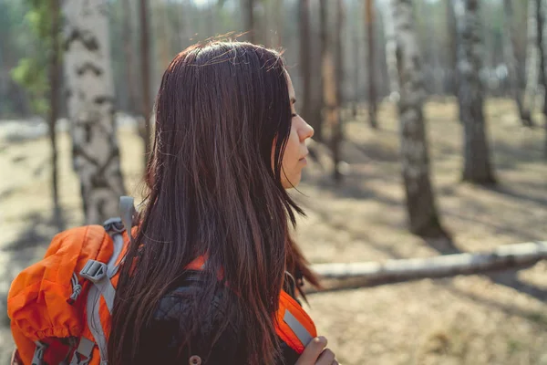 Traveling woman with backpack in woodsSide view of brunette standing with bright orange backpack in tranquil sunny forest looking away