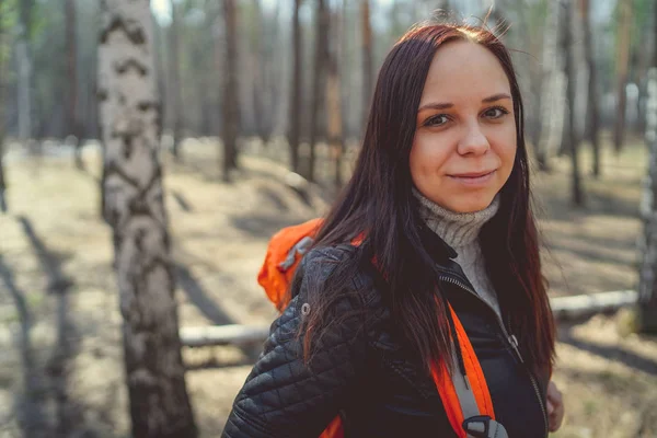 Traveling woman with backpack in woodsSide view of brunette standing with bright orange backpack in tranquil sunny forest looking away