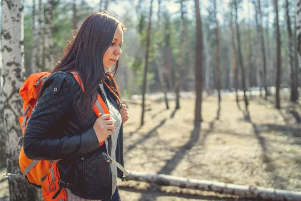 Traveling woman with backpack in woodsSide view of brunette standing with bright orange backpack in tranquil sunny forest looking away