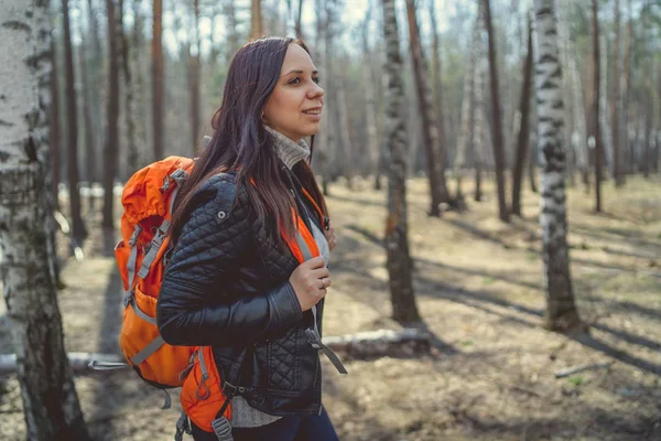 Traveling woman with backpack in woodsSide view of brunette standing with bright orange backpack in tranquil sunny forest looking away