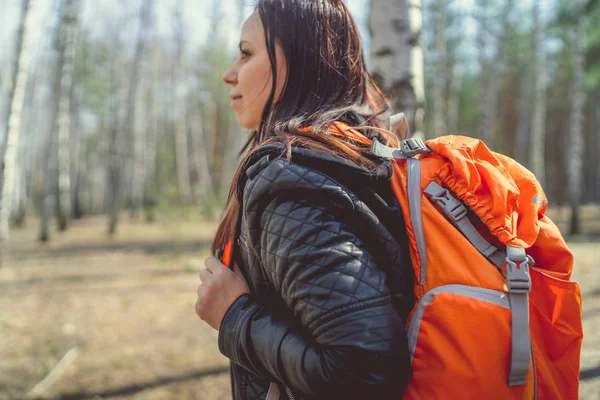 Traveling woman with backpack in woodsSide view of brunette standing with bright orange backpack in tranquil sunny forest looking away