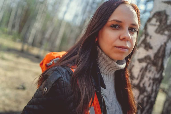 Traveling woman with backpack in woods Side view of brunette standing with bright orange backpack in tranquil sunny forest looking away