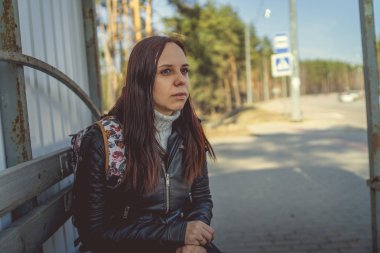 Otobüs durağında oturan esmer, siyah deri ceketli sıradan bir kadın otobüs durağında oturmuş, başka yöne bakan toplu taşıma araçlarını bekliyor.