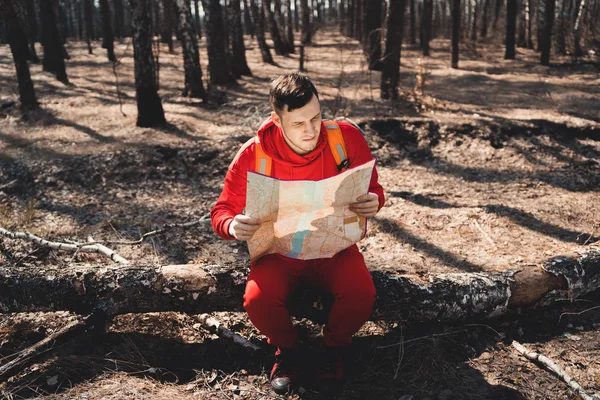 Man with backpack reading map in woodsYoung inspired man in red sportswear sitting on fallen tree trunk in forest and reading map while traveling with backpack