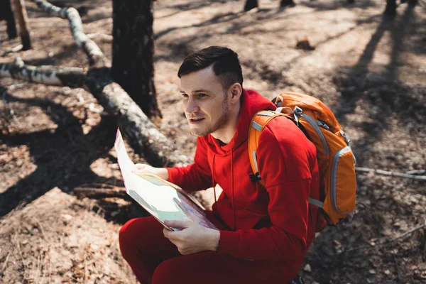 Man with backpack reading map in woodsYoung inspired man in red sportswear sitting on fallen tree trunk in forest and reading map while traveling with backpack