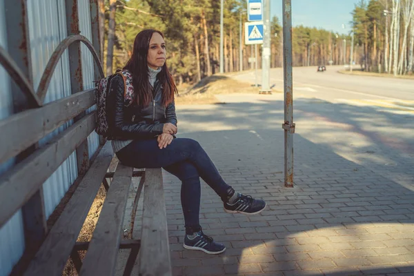 年轻的黑发女子坐在公共汽车站上 身穿黑色皮大衣的妇女坐在公共汽车站上等公共交通工具 望着远方 — 图库照片