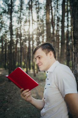 Ormanda kitap okuyan bir adam, bir ağacın yanında duruyor. Beyaz gömlekli mutlu bir erkek güneşli ormanda kitap okuyor.