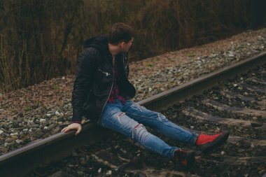 Rayların üzerinde oturan modaya uygun genç adam. Kotlu ve deri ceketli dalgın bir erkek bulutlu bir günde tren yolunda oturuyor.