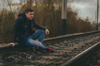 Rayların üzerinde oturan modaya uygun genç adam. Kotlu ve deri ceketli dalgın bir erkek bulutlu bir günde tren yolunda oturuyor.