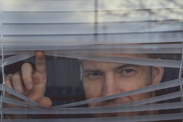 Man watching through window blinds. Portrait of young thoughtful male with brown eyes observing through window jalousie