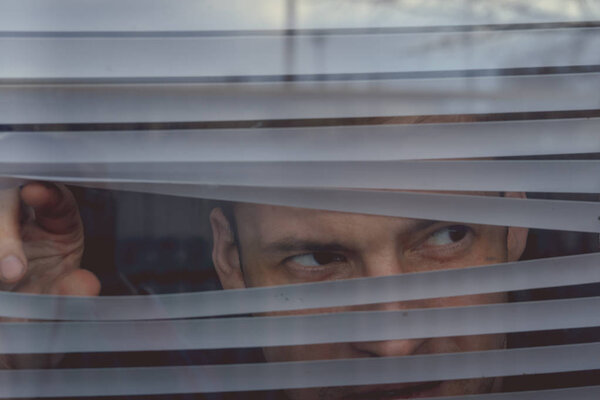 Man watching through window blinds. Portrait of young thoughtful male with brown eyes observing through window jalousie