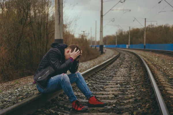 Demiryolunda oturan umutsuz adam. Sonbahar kırsalında raylarda otururken kafasına dokunan sıradan giysili umutsuz bir erkeğin yan görüntüsü