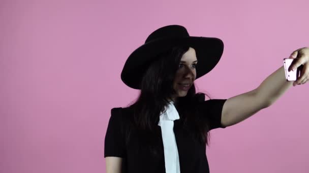 La dama toma una selfie sobre un fondo rosa. Una mujer con un vestido negro y un sombrero toma una foto de sí misma — Vídeo de stock