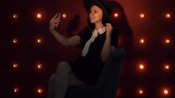 Signora seduta su una sedia e si fa un selfie su smartphone. Una bella donna vestita di nero che si fotografa al telefono, su uno sfondo rosso . — Video Stock